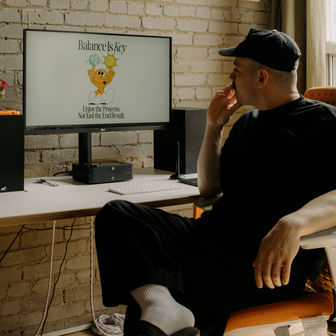 A person wearing a black cap sits at a desk and looks at a computer monitor.