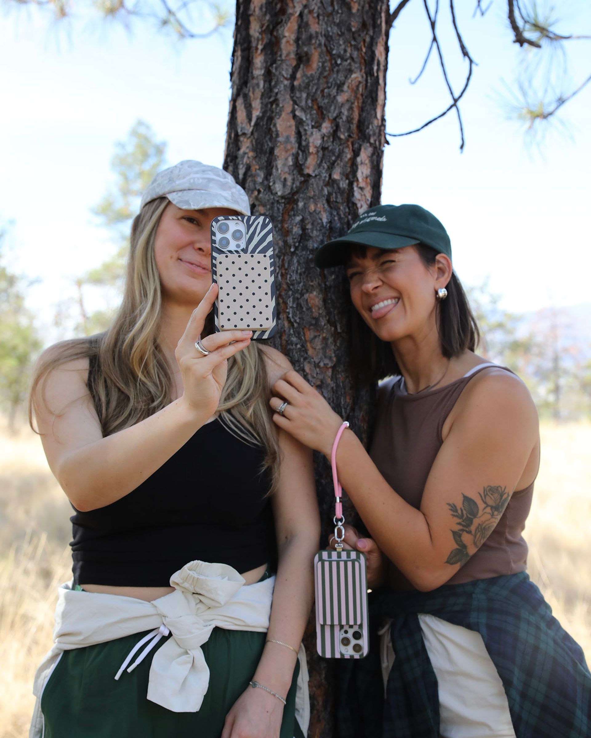 Two women in baseball caps take a playful selfie together outdoors by a tree.