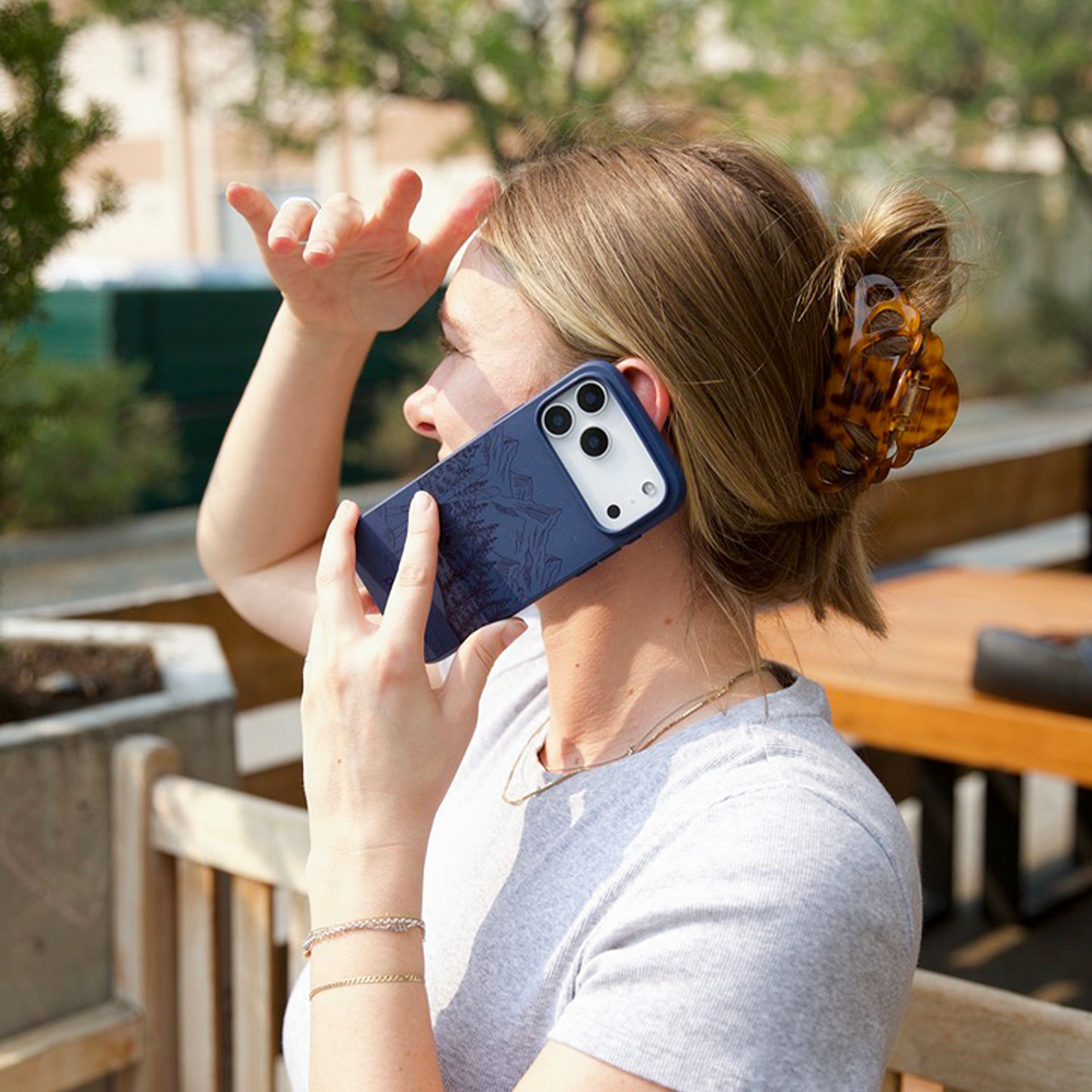 A woman with her hair in a claw clip talks on a blue-cased smartphone while outdoors.