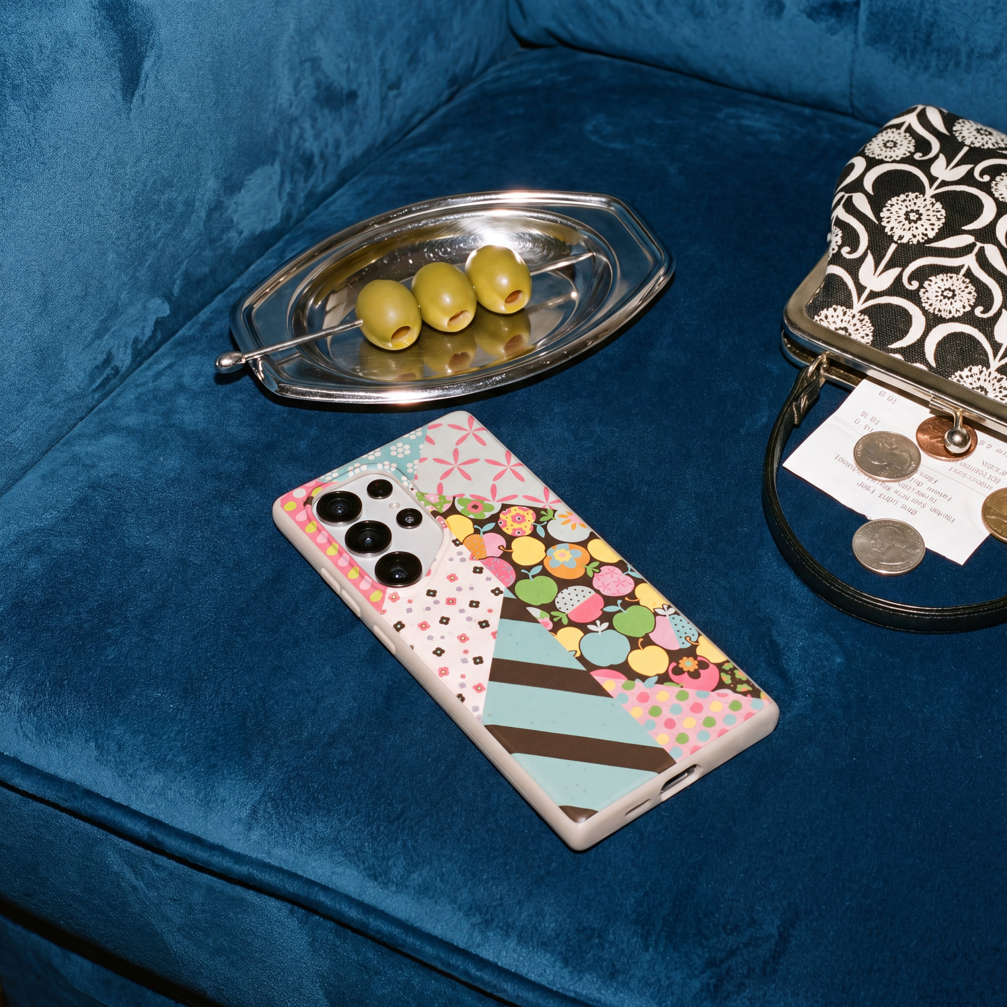 A phone in a colorful case, a dish of olives, and a purse on a blue velvet couch.