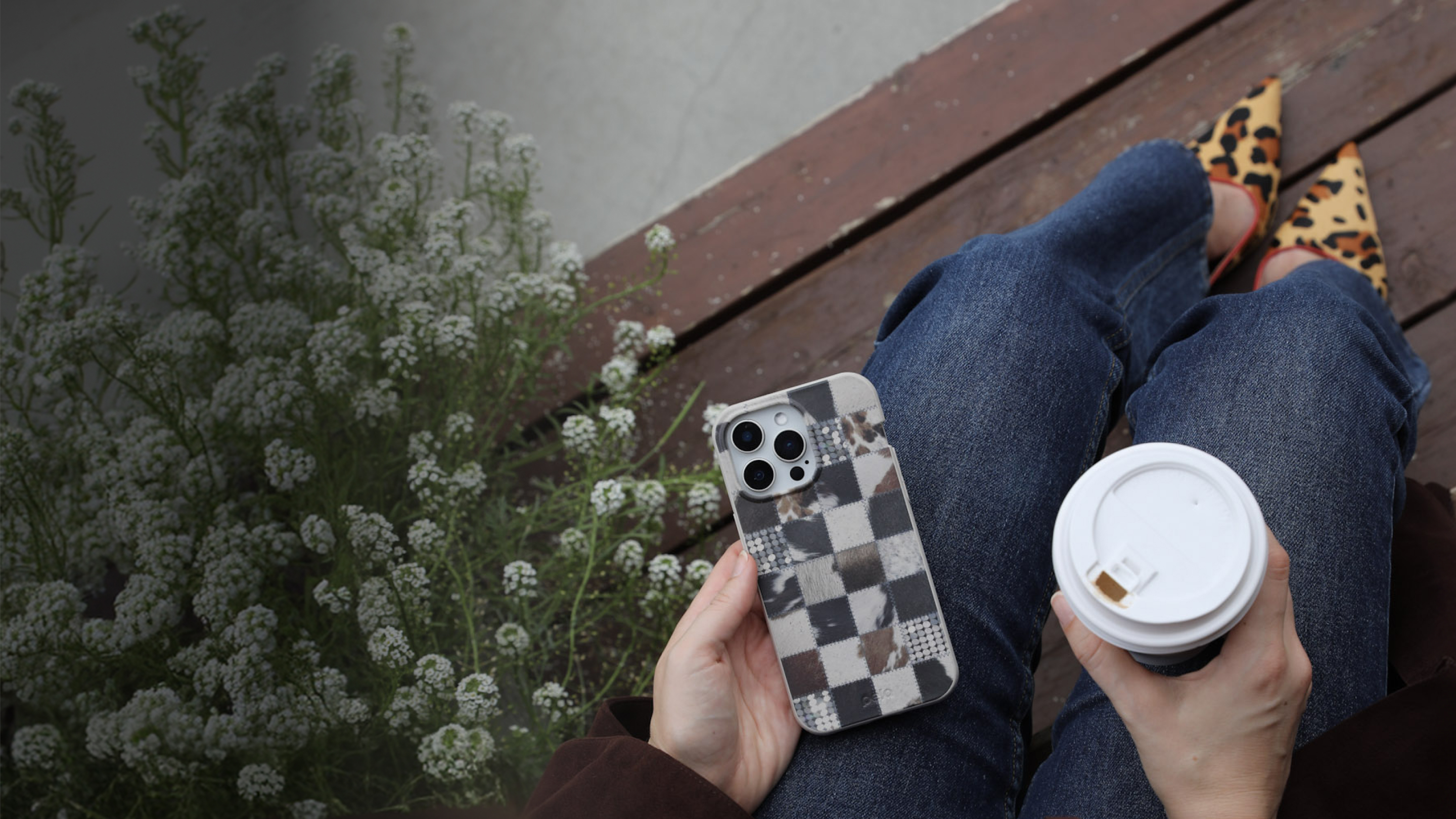 A person holding a phone in a patchwork case and a coffee cup, sitting by white flowers.