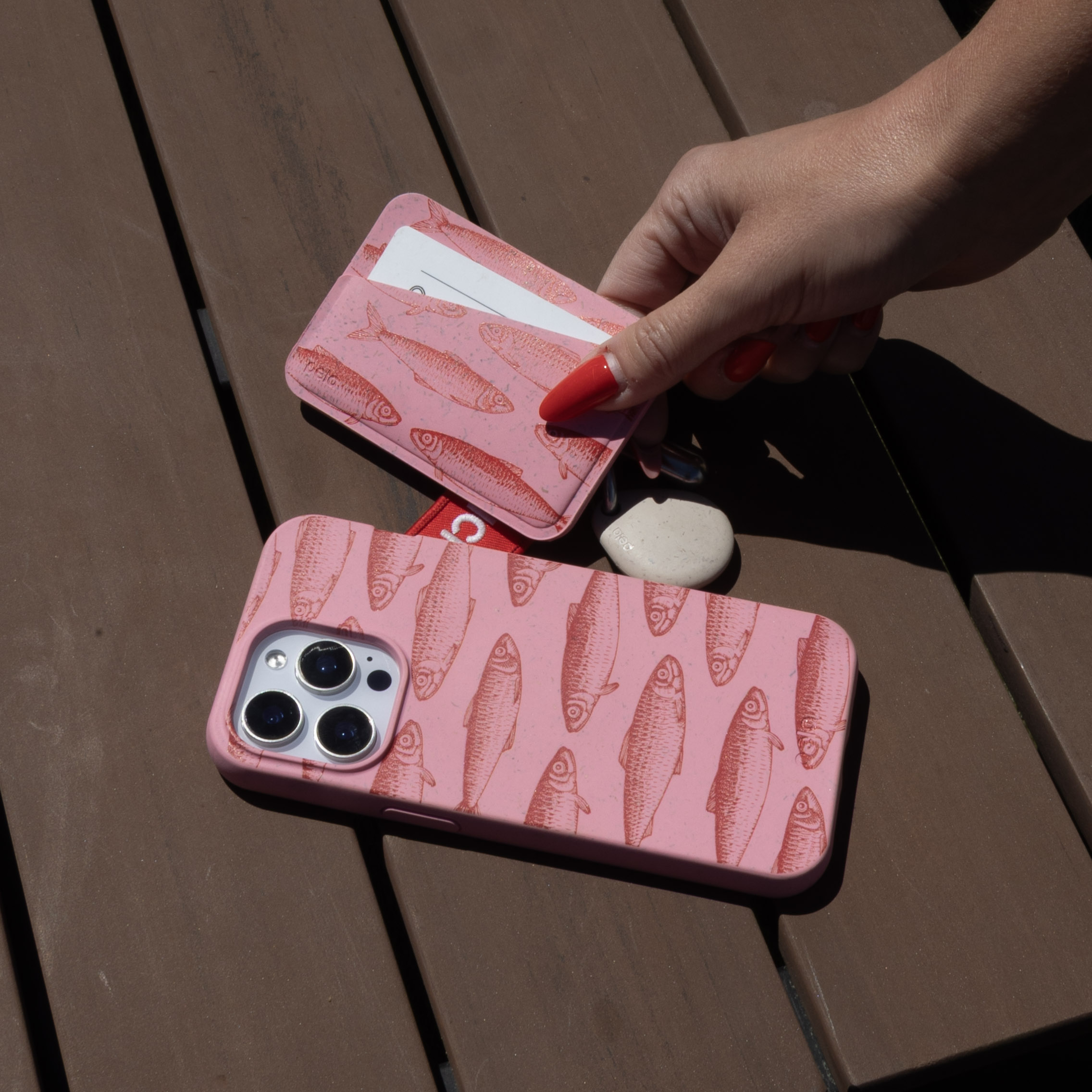 A hand holding a pink, fish-patterned card wallet over a matching phone case on a wooden surface.