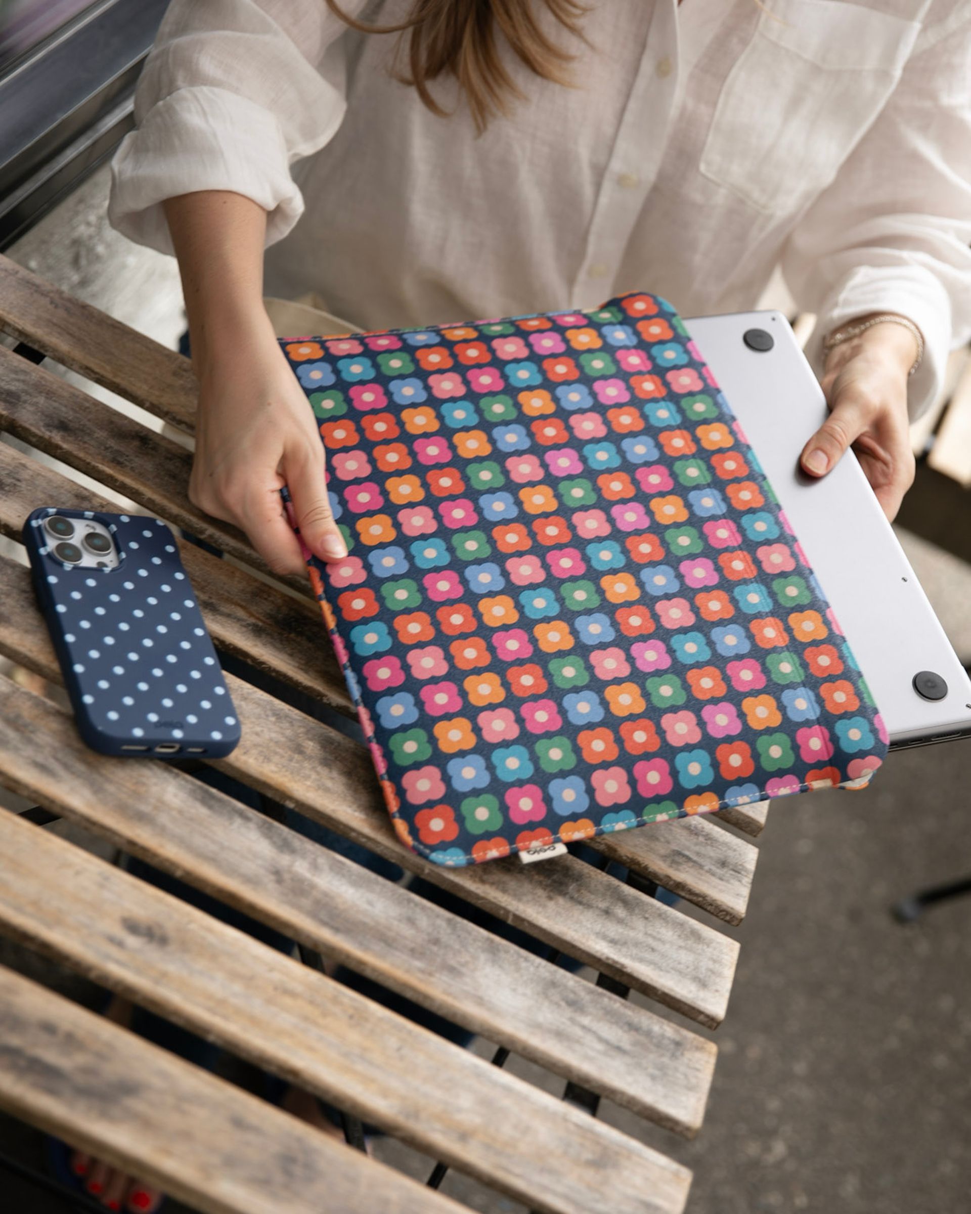 A person slides a laptop into a colorful floral sleeve, with a polka-dot phone case nearby.