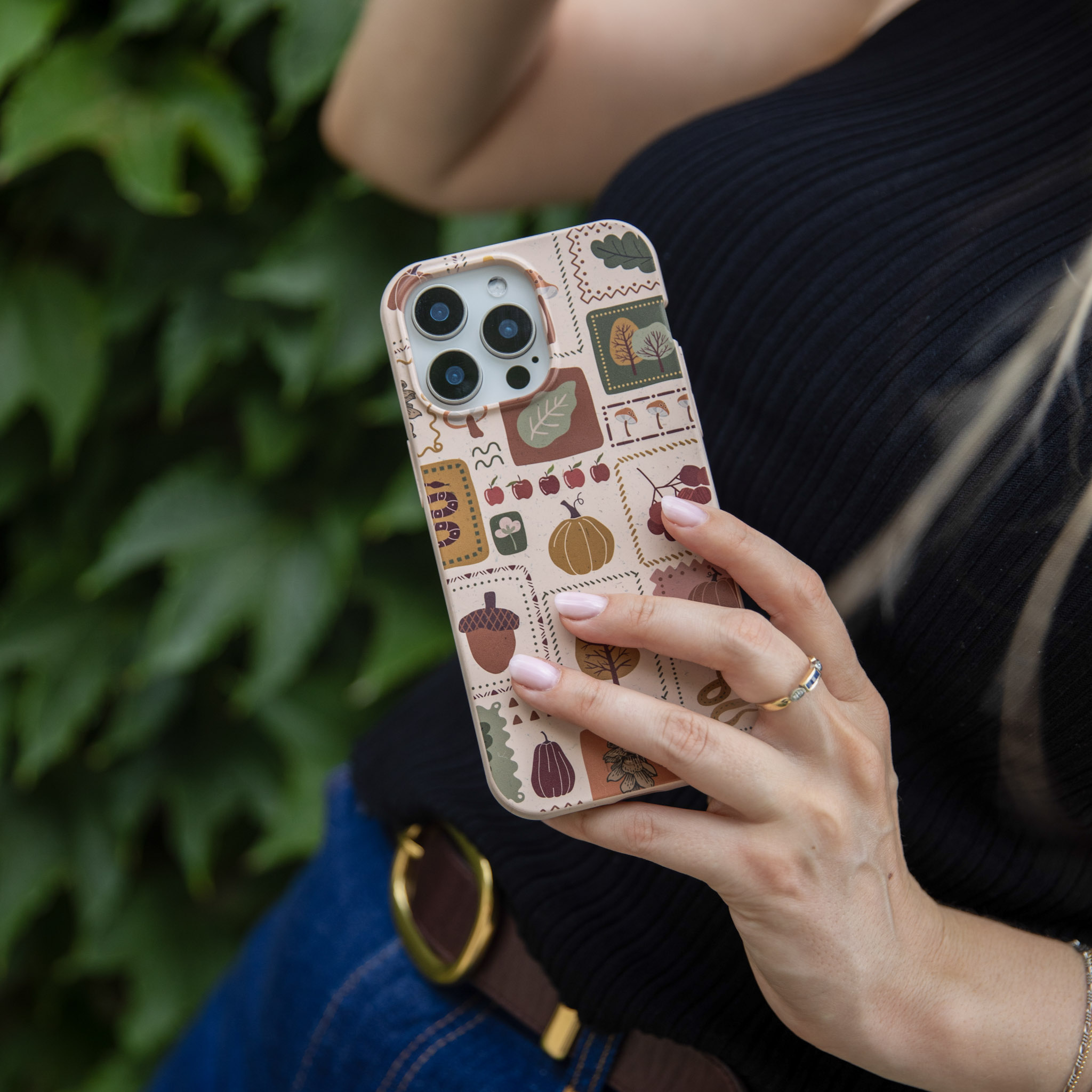 A person holding a phone with a beige, autumn-themed patterned case in front of green ivy.