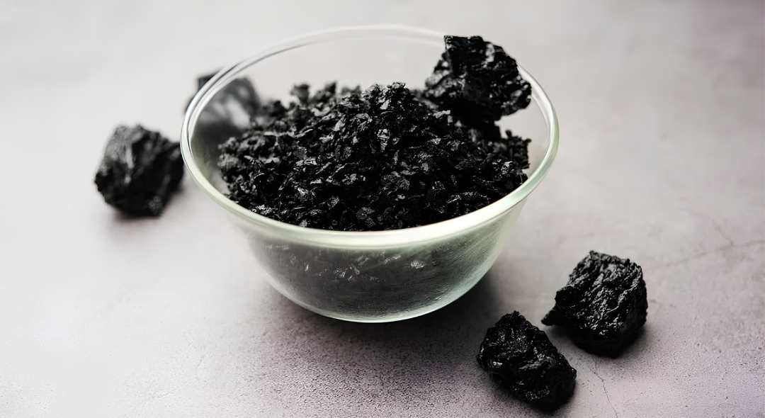 Glass bowl with black flaky sea salt on a gray surface.