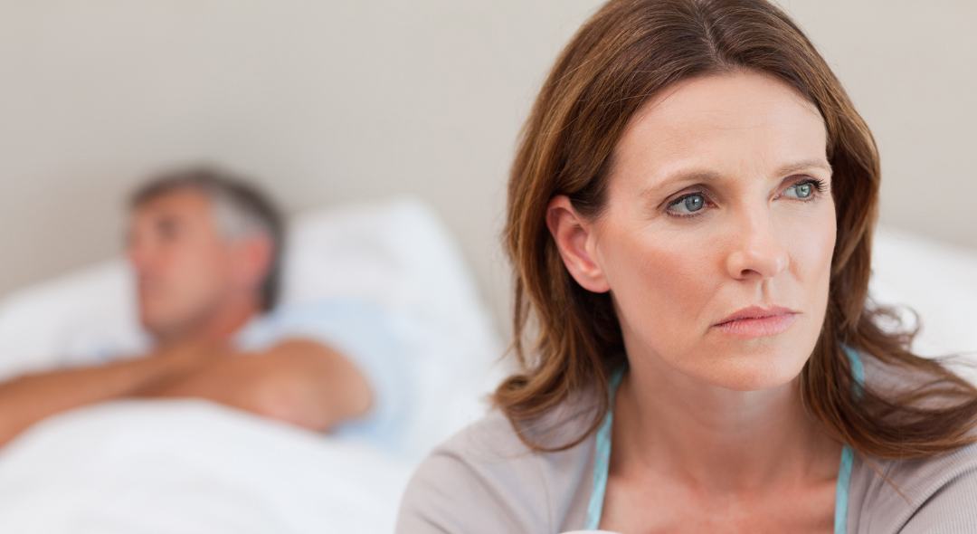 Woman looking pensive, with a man lying behind her in bed.