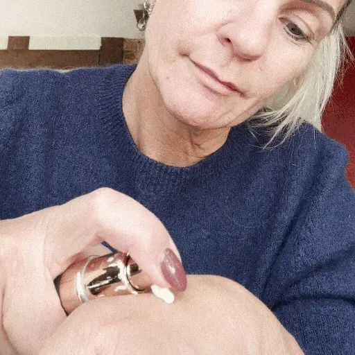 A woman in a blue sweater pumps a small amount of white cream onto the back of her hand.