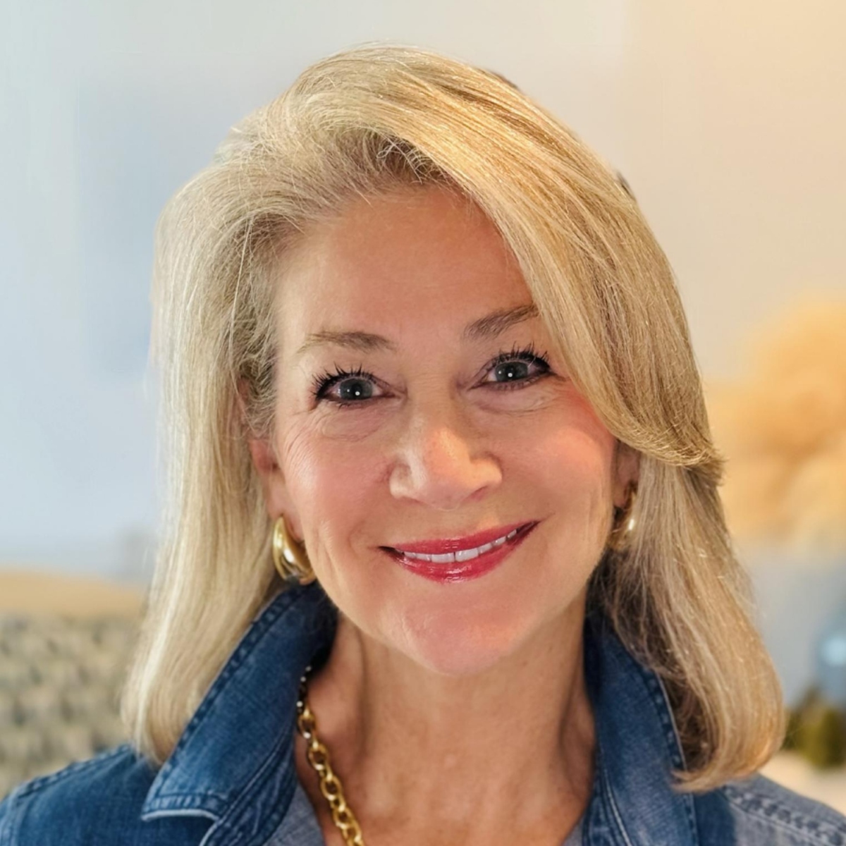 A smiling woman with blonde hair, wearing a denim jacket, gold earrings, and a gold chain necklace.