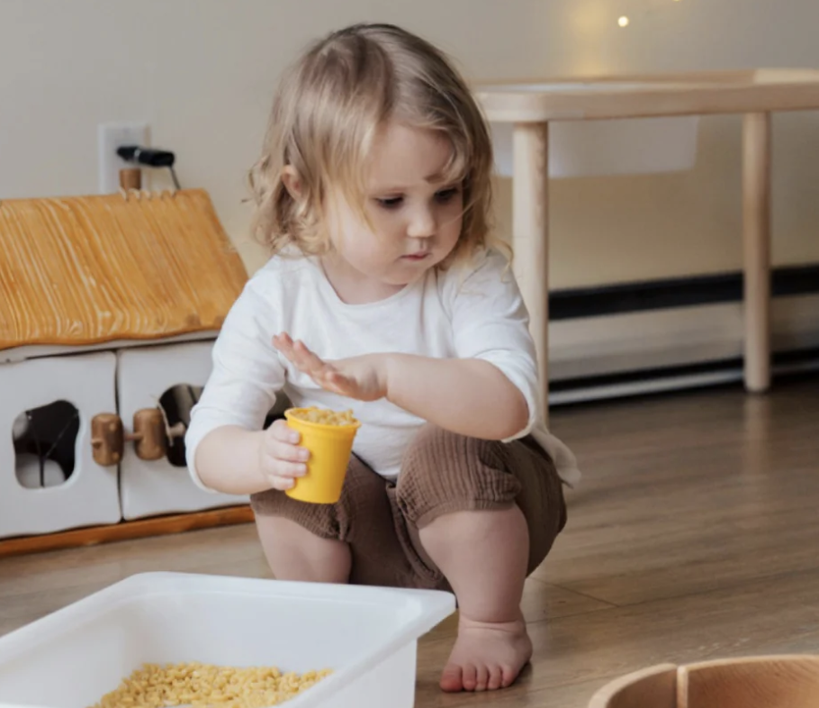 Child playing with a yellow cup and pasta in a room with wooden toys.