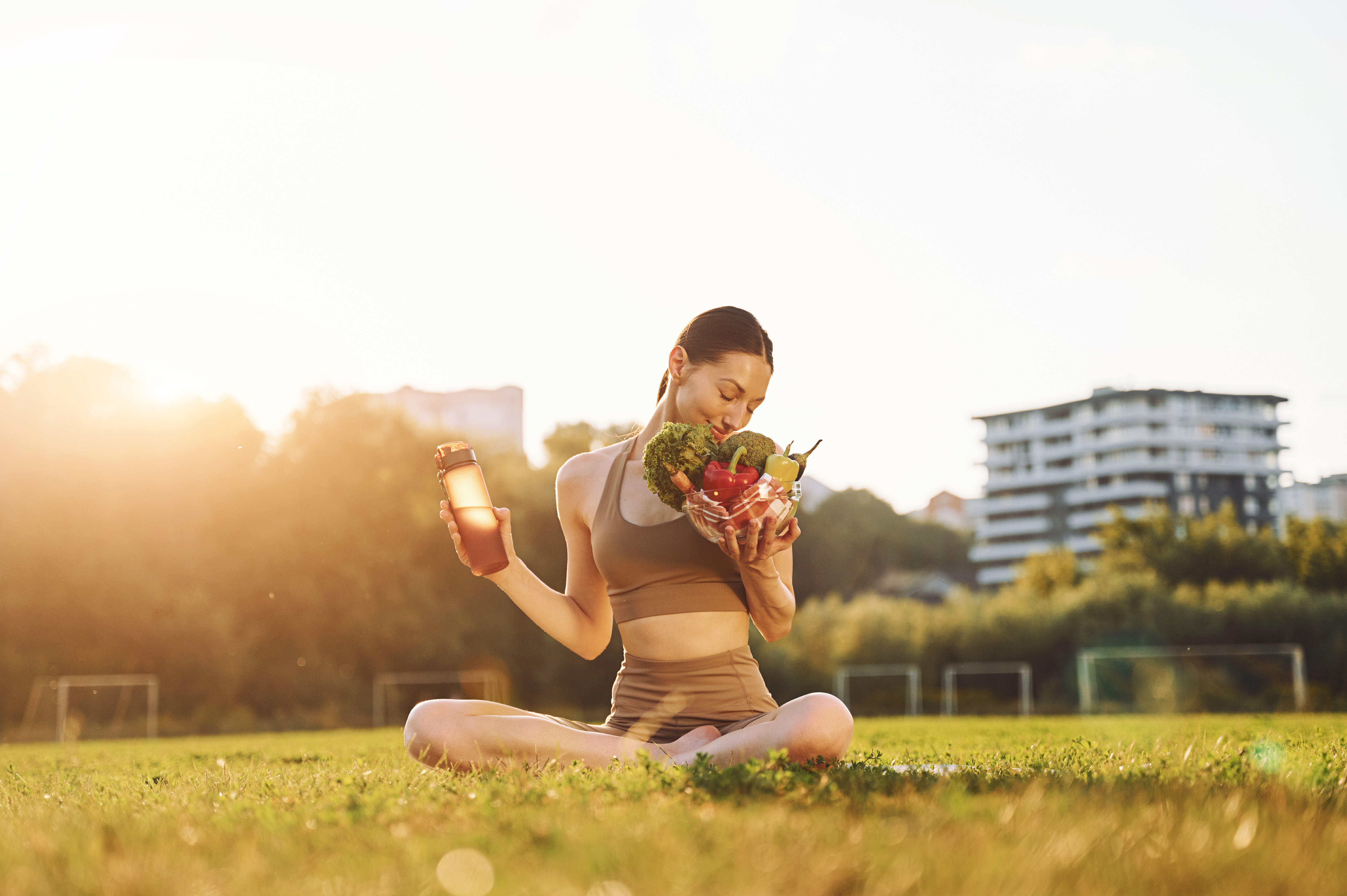Person sitting on grass holding a bowl of vegetables and a drink bottle.