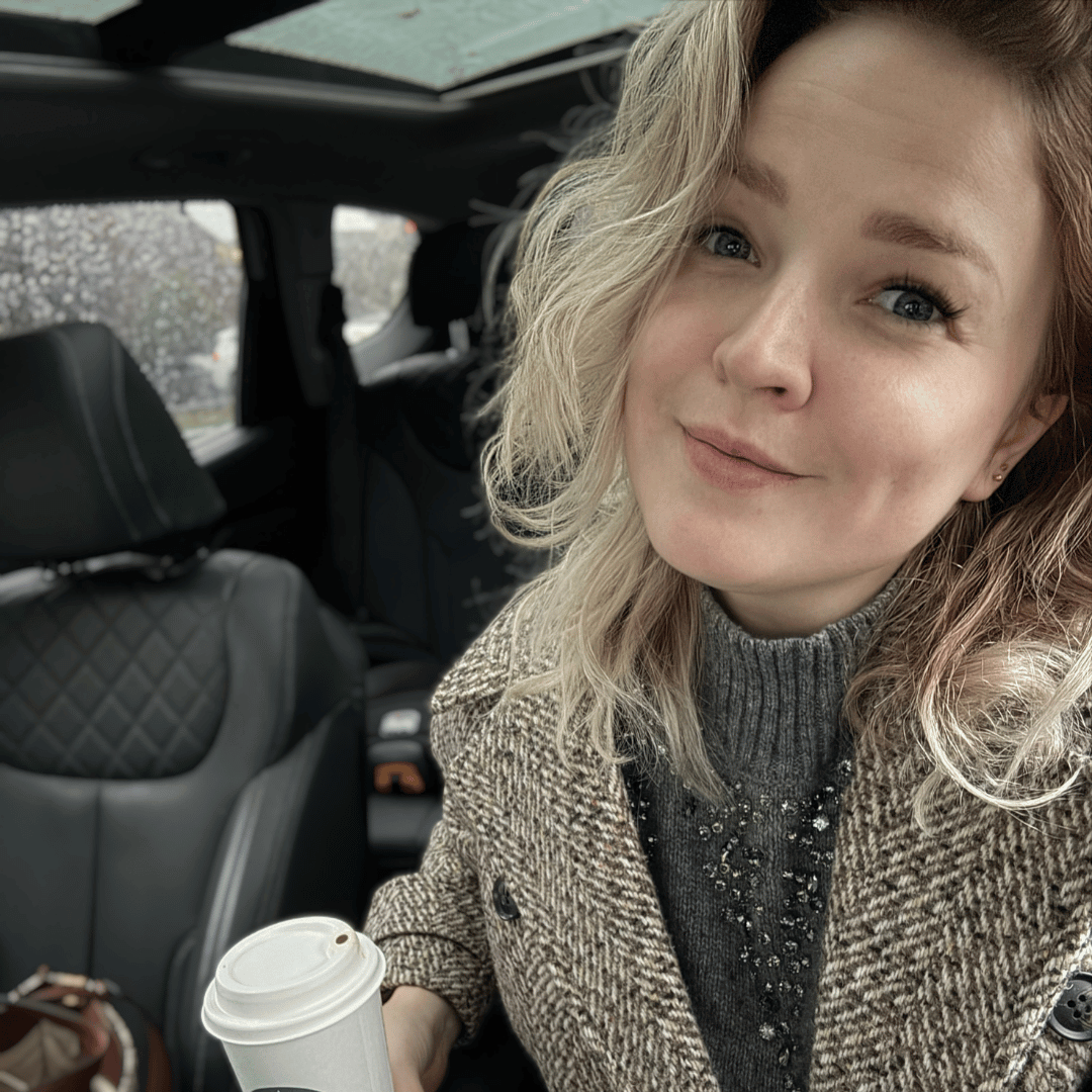 A woman with blonde hair smiles while sitting in a car, holding a disposable coffee cup.