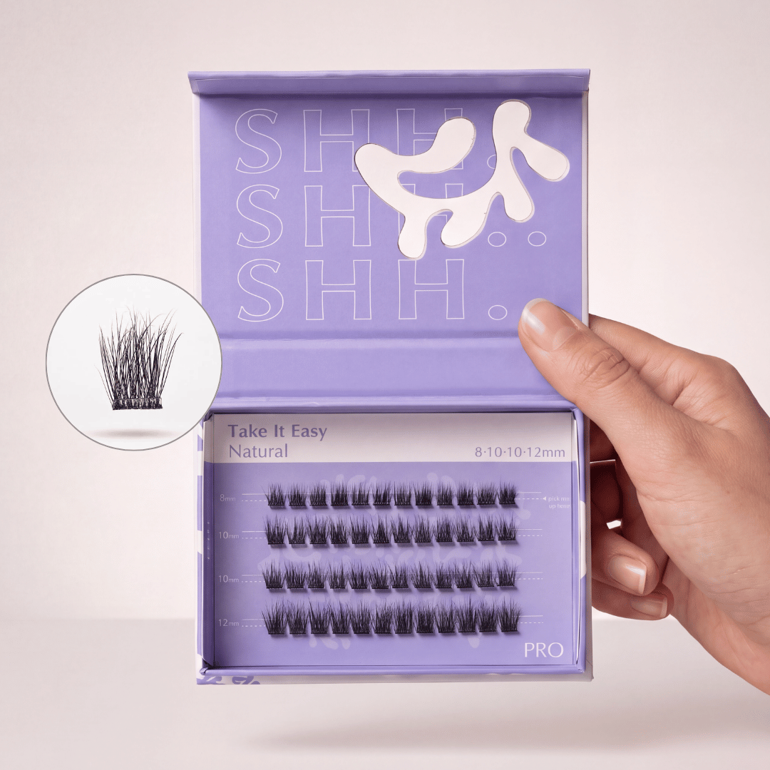A hand holding an open purple box of 'Take It Easy Natural' false eyelashes of varying lengths.