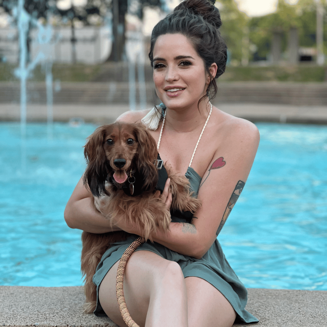 A woman holds a small, brown, long-haired dog while sitting in front of a fountain.