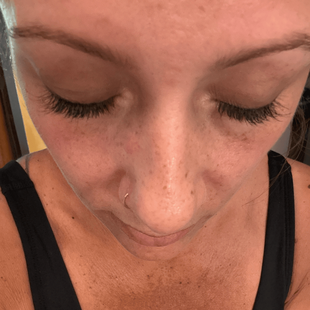A close-up, high-angle view of a woman with closed eyes, showing long eyelash extensions and a nose ring.