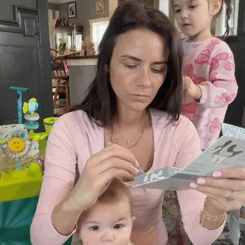 A woman applying makeup with a baby in front of her and a toddler standing behind her.