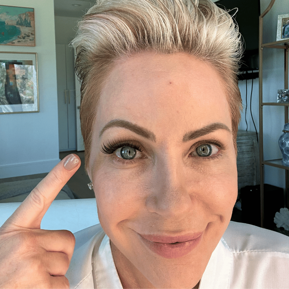 Person with short hair pointing to their eye, wearing makeup.