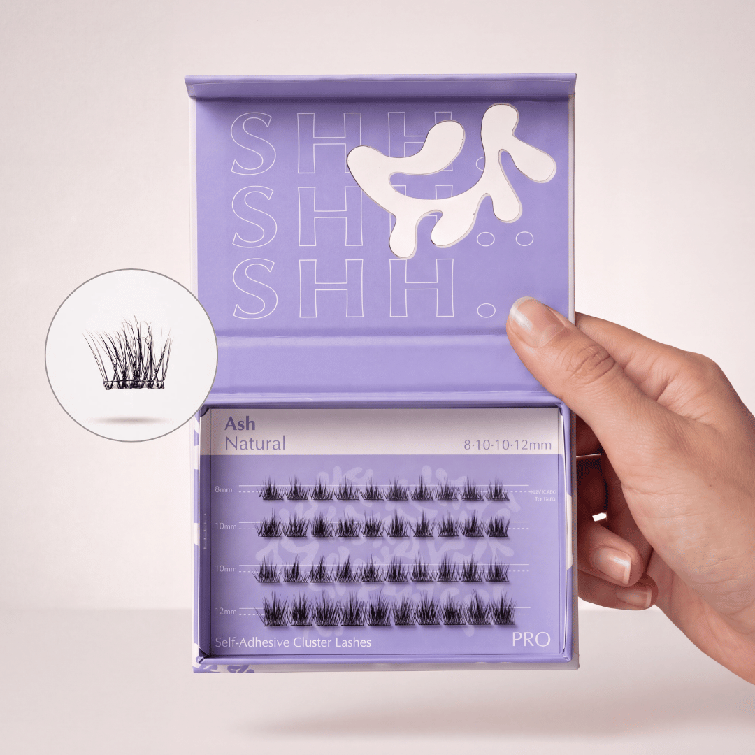 A hand holds open a purple box of Ash Natural self-adhesive cluster lashes.