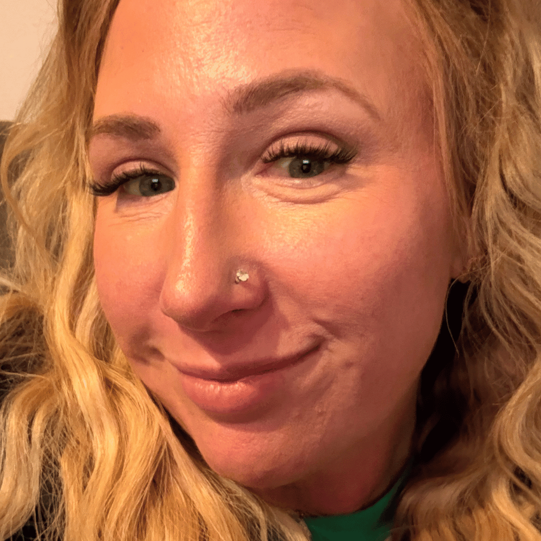 A close-up photo of a smiling woman with blonde, curly hair and a nose piercing.