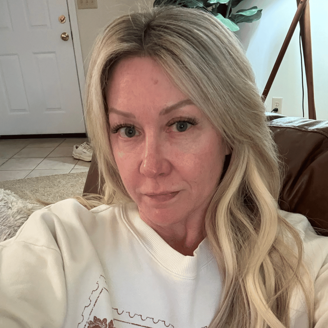 A woman with long blonde hair wearing a cream sweatshirt looks at the camera while sitting indoors.