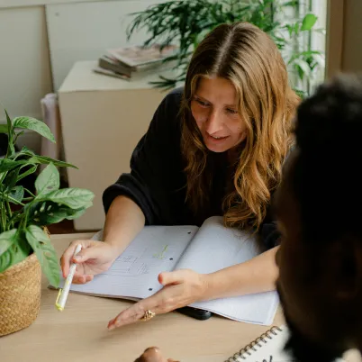 Two people engaged in a discussion at a table with plants and notebooks.
