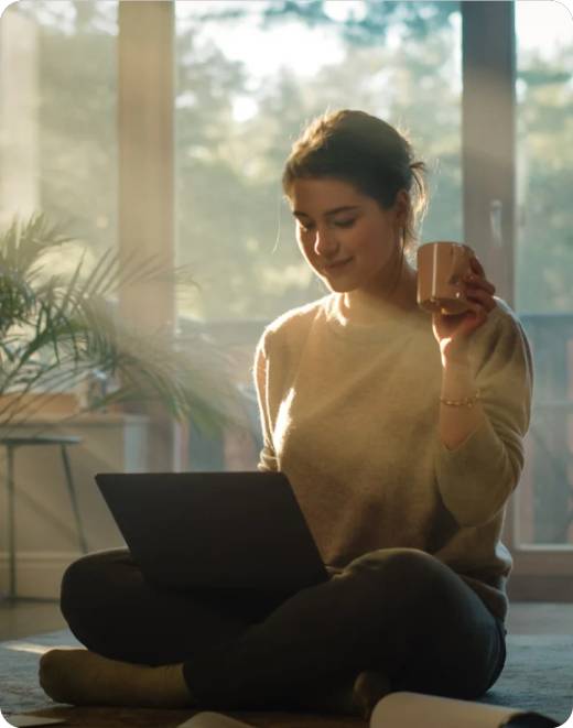 Person sitting with a laptop and holding a mug in a sunlit room.