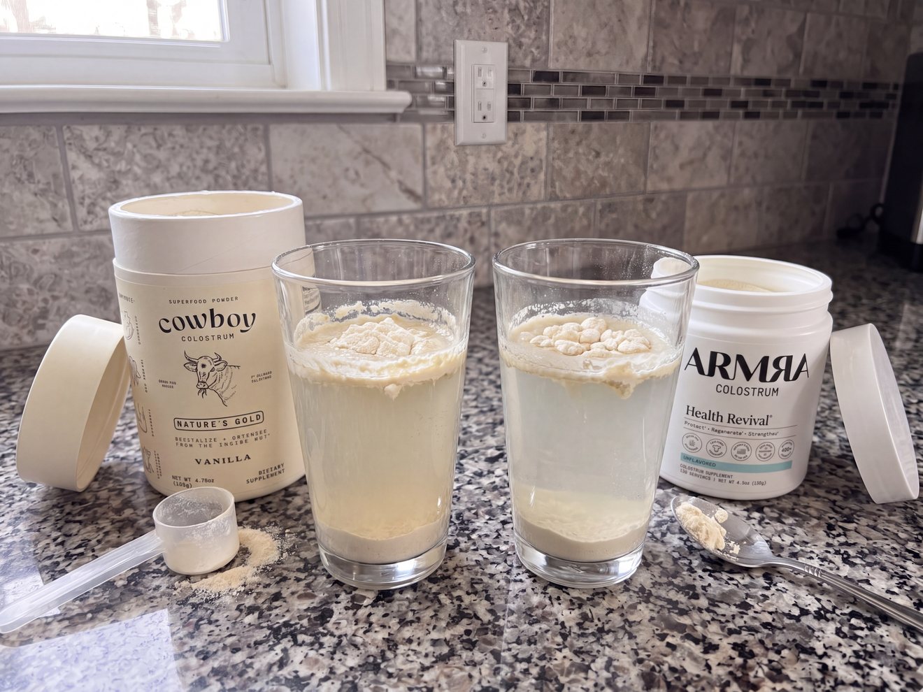 Two brands of colostrum powder, Cowboy and ARMRA, are mixed into two glasses of water on a countertop.