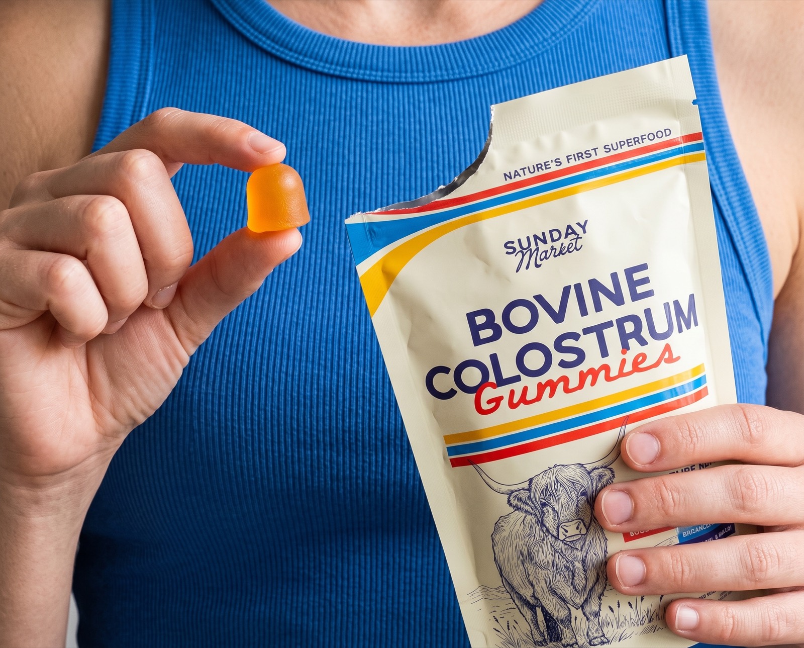 A person in a blue top holds a single orange gummy and a pouch of Sunday Market Bovine Colostrum Gummies.