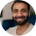A circular, slightly blurry headshot of a smiling man with a beard and dark hair.