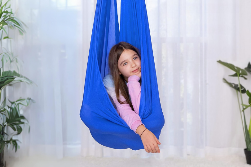 Child in a blue hammock swing indoors, surrounded by plants.