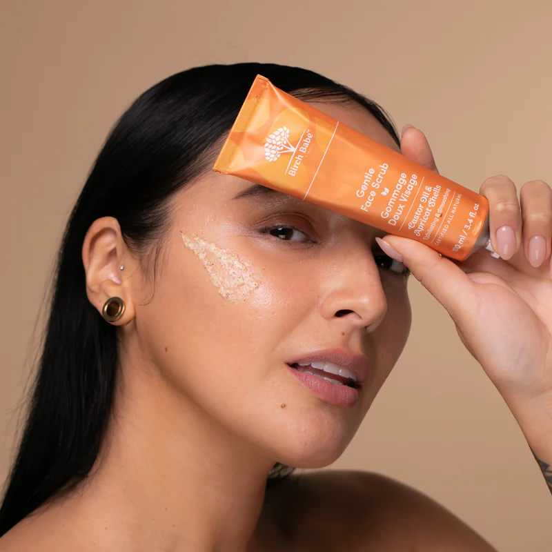 Person holding an orange face scrub tube near their face with product applied.