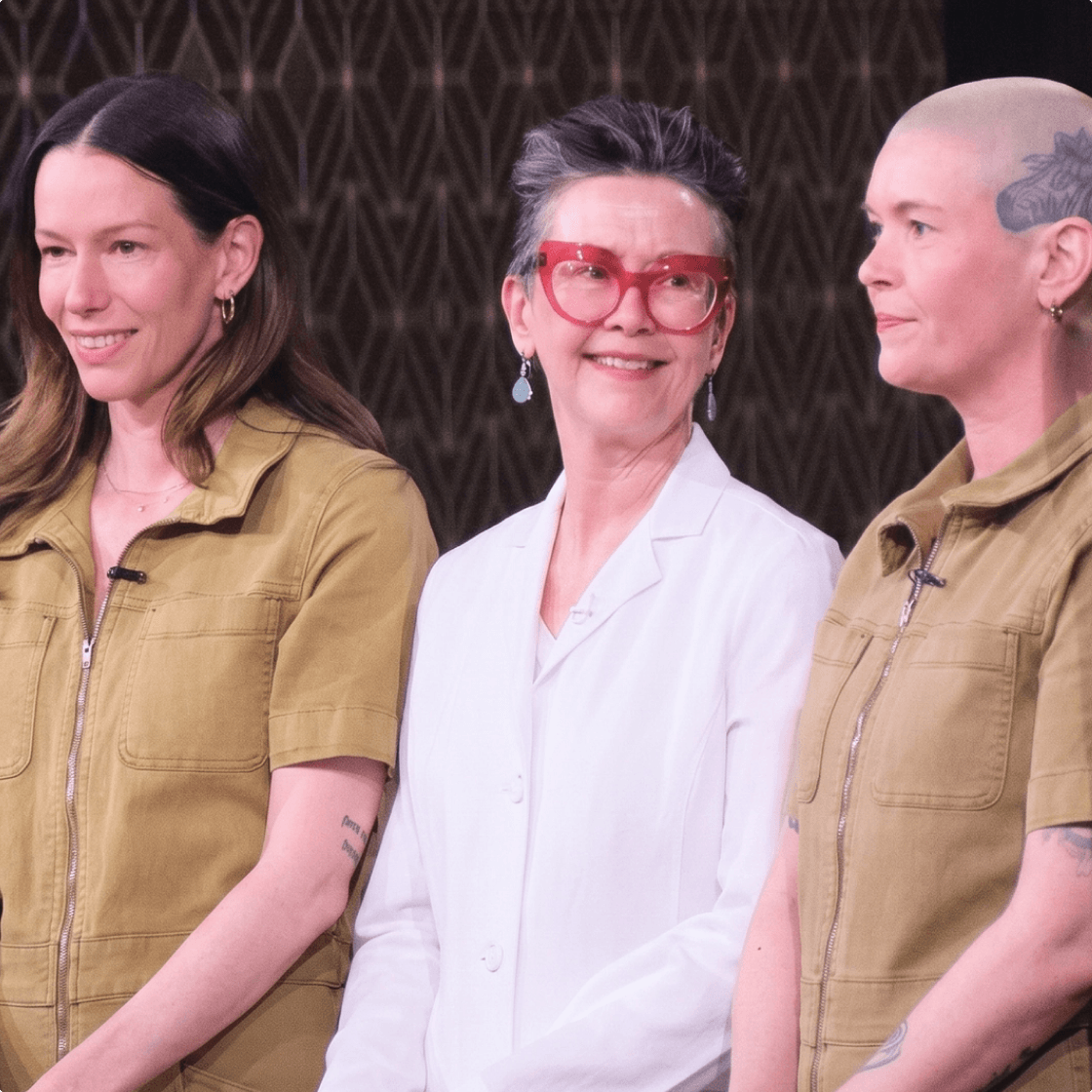 Three women standing together; one in a white jacket and red glasses between two women in green jumpsuits.