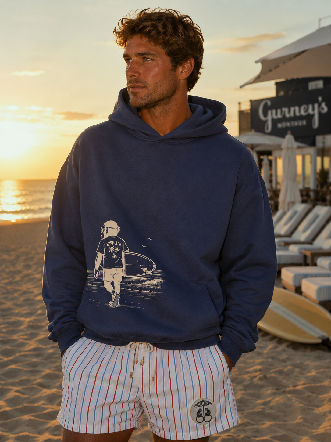 A man in a blue hoodie and striped shorts stands on a beach at sunset.