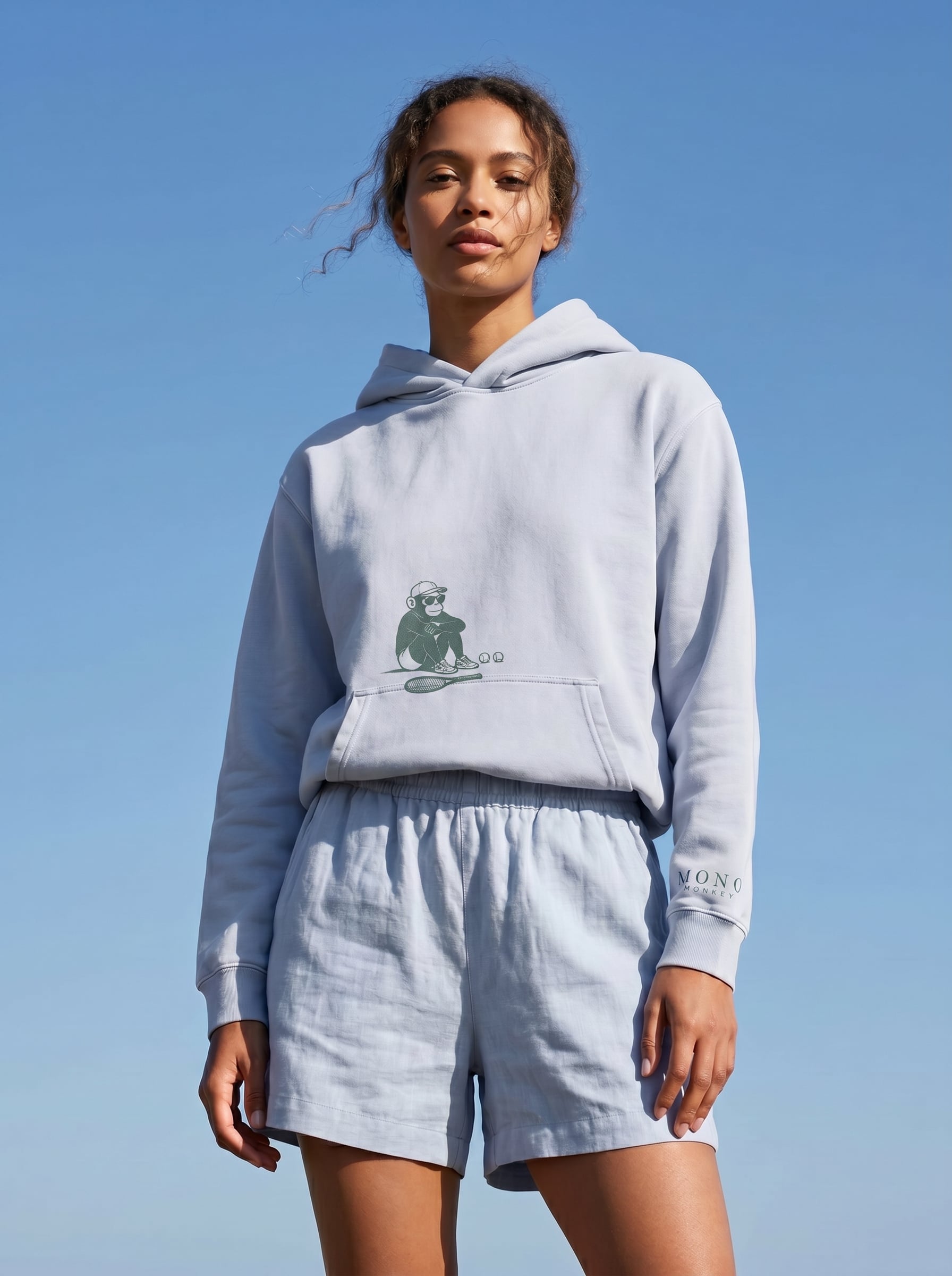 A woman in a light blue hoodie and shorts with a tennis monkey graphic against a blue sky.