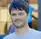 A low-resolution, pixelated headshot of a man with dark hair against a blurry background.