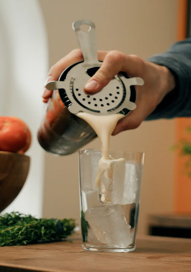Hand pouring a drink through a strainer into a glass with ice, apples in the background.