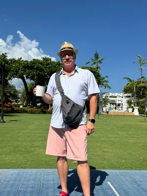 Man in hat and shorts holds mug, standing outdoors on a sunny day.