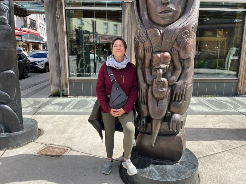 Person standing next to a carved wooden totem pole in an urban setting.