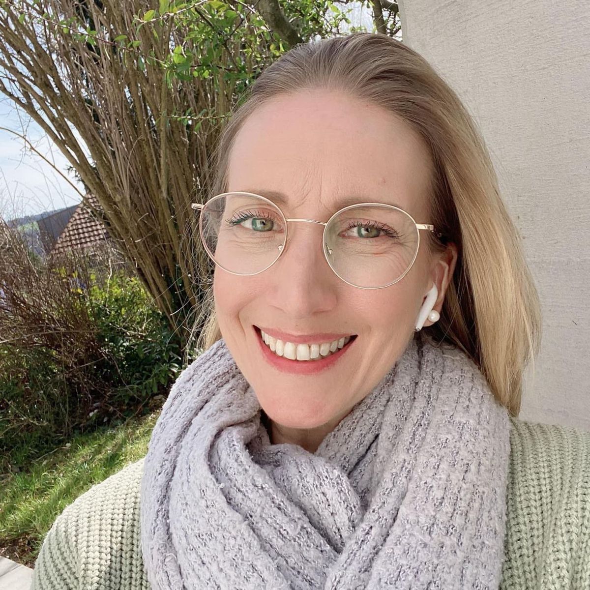 Person smiling, wearing glasses and earphones, with a scarf in an outdoor setting.