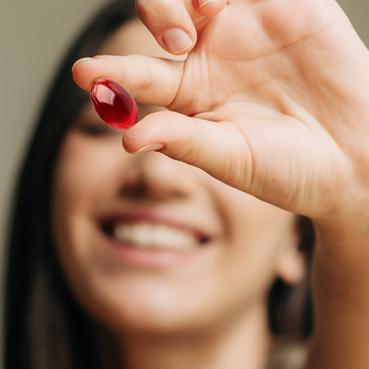 Person holding a red capsule between fingers, smiling in the background.