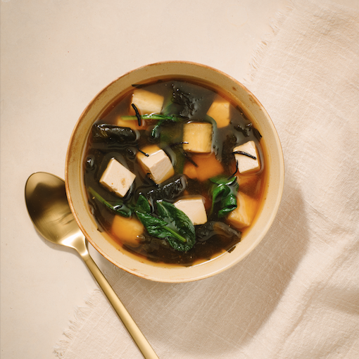 Bowl of miso soup with tofu, seaweed, and spinach, next to a gold spoon.