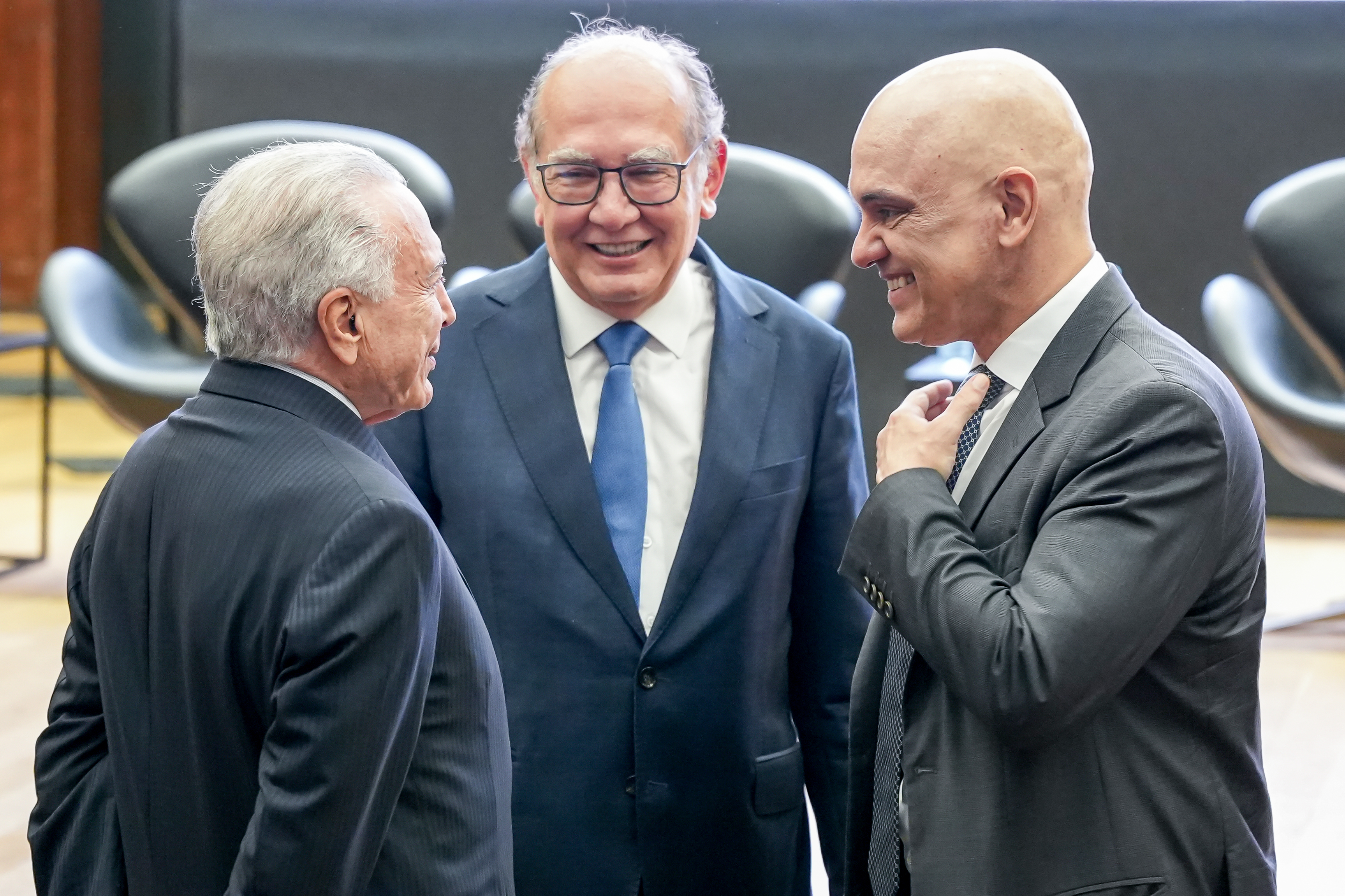 Three men in suits are standing together indoors, smiling and talking with each other.