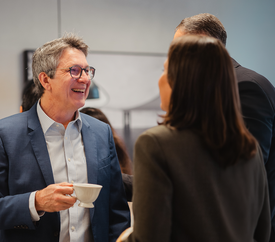 A smiling man in a blue blazer and glasses holds a teacup while talking to two people.