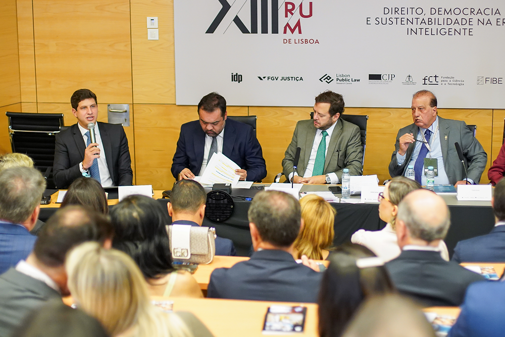 A panel of four men in suits sits at a table, one speaking to an audience.
