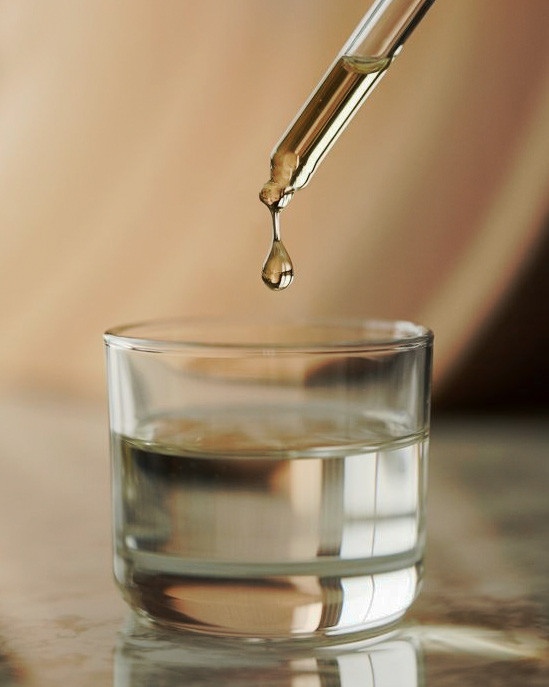 A close-up of a drop of golden liquid falling from a glass dropper into a glass of water.