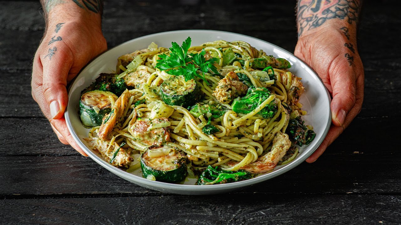 Hands holding a plate of pasta with vegetables and garnish.
