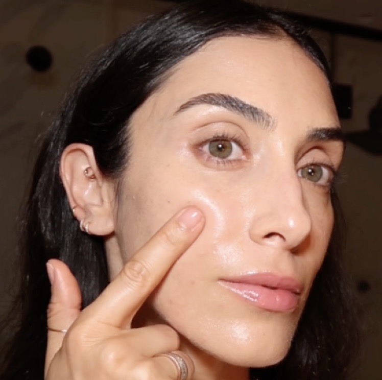 A close-up of a woman with dark hair and glowing skin touching her cheek with her finger.