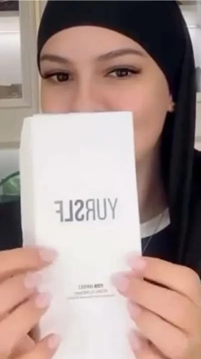 A woman in a black head covering holds up a white box with the word 'YURSLEF' on it.
