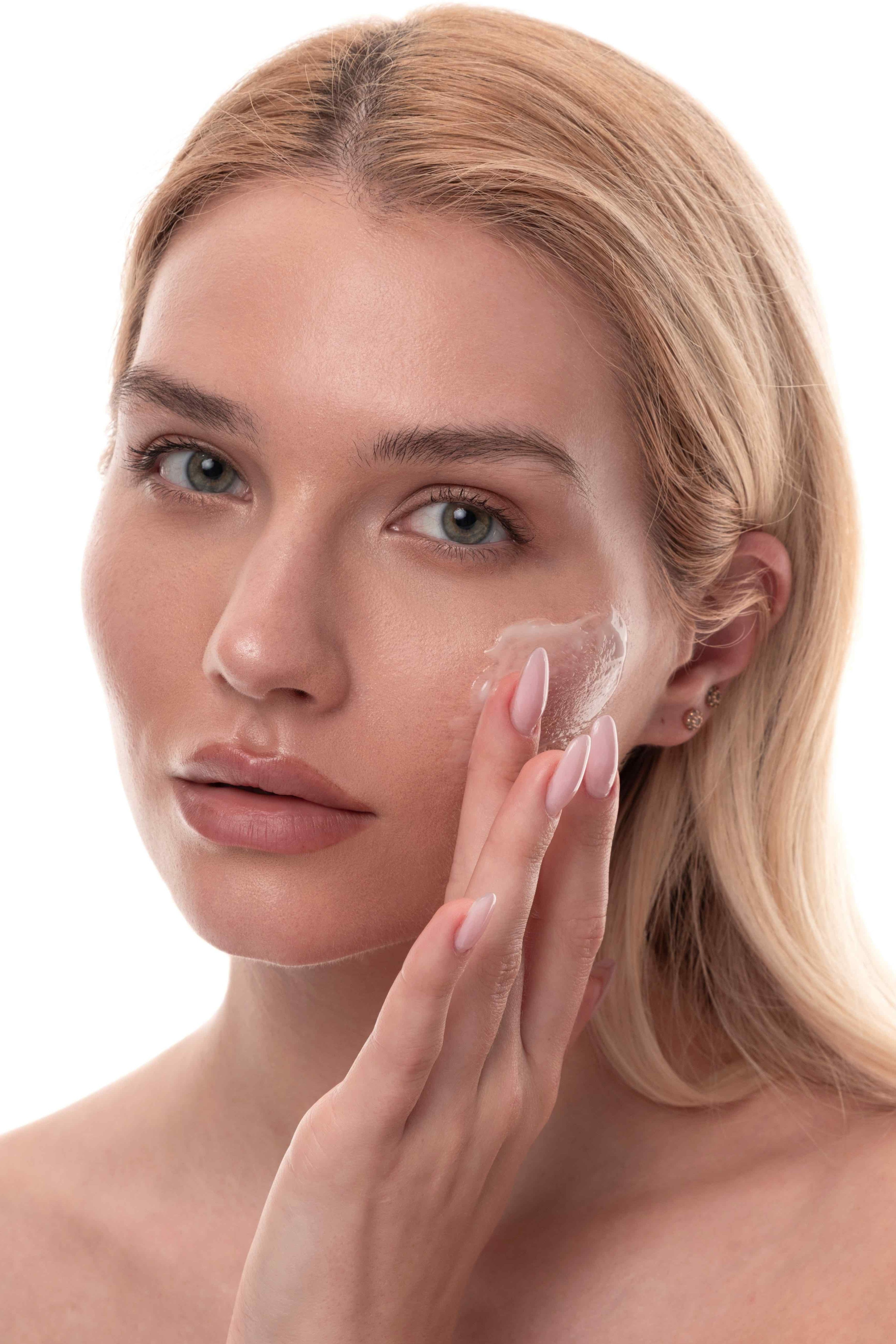 A close-up of a blonde woman with green eyes applying a clear gel to her cheek.