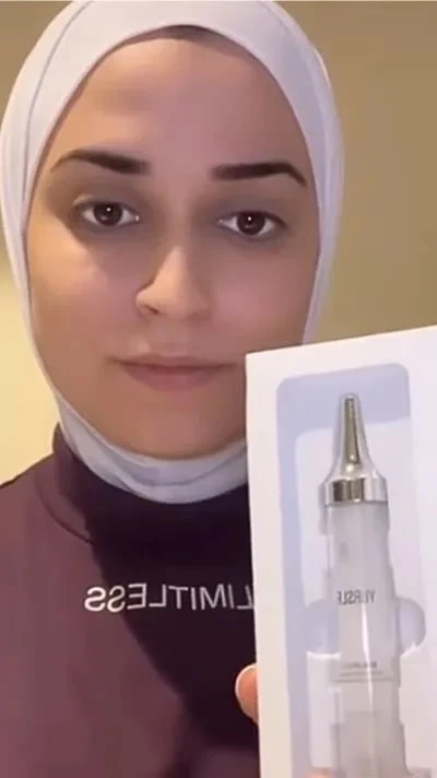 A person in a white hijab and purple shirt holds up a box containing a cosmetic applicator bottle.