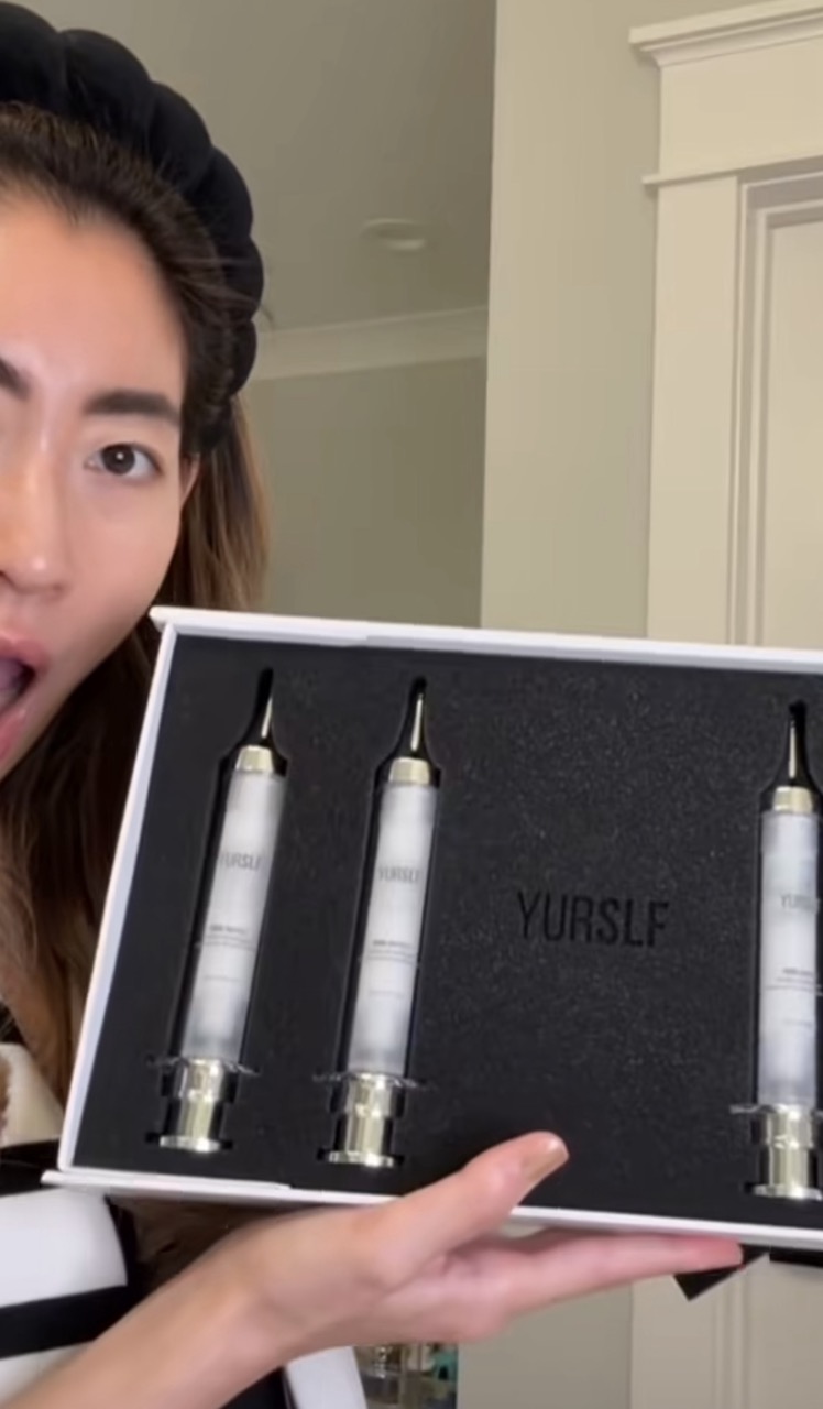 A woman with a surprised expression holds an open box of YURSLF cosmetic applicators shaped like syringes.